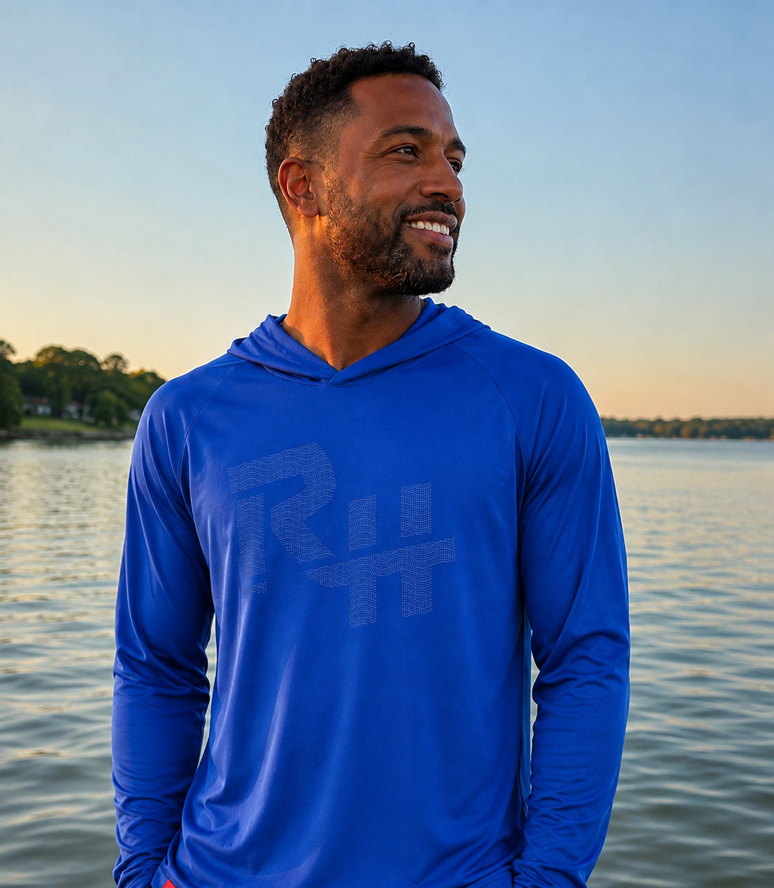A man wearing a royal blue long-sleeve hooded shirt with a tonal gray 'RH' logo, standing outdoors by a calm lake at sunset.