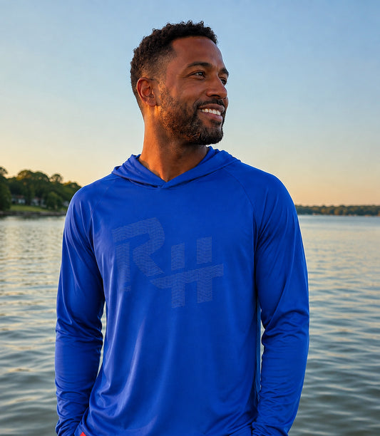 A man wearing a royal blue long-sleeve hooded shirt with a tonal gray 'RH' logo, standing outdoors by a calm lake at sunset.